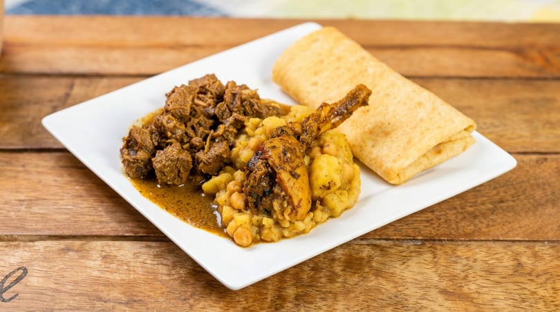 A white plate with stewed meat, chicken, chickpeas, potatoes, and a folded flatbread.