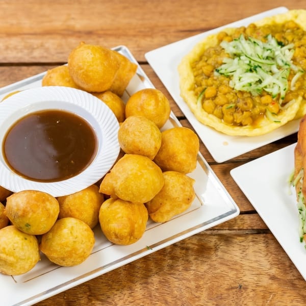 A wooden table holds three plates of Caribbean food: fried dough balls with sauce, curried chickpeas, and fried bread with cucumber.