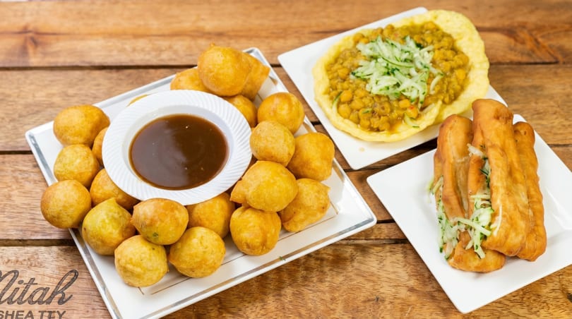 A wooden table holds three plates of Caribbean food: fried dough balls with sauce, curried chickpeas, and fried bread with cucumber.