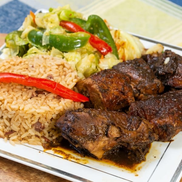 A plate of brown stew chicken, rice and peas, and steamed cabbage with red peppers.